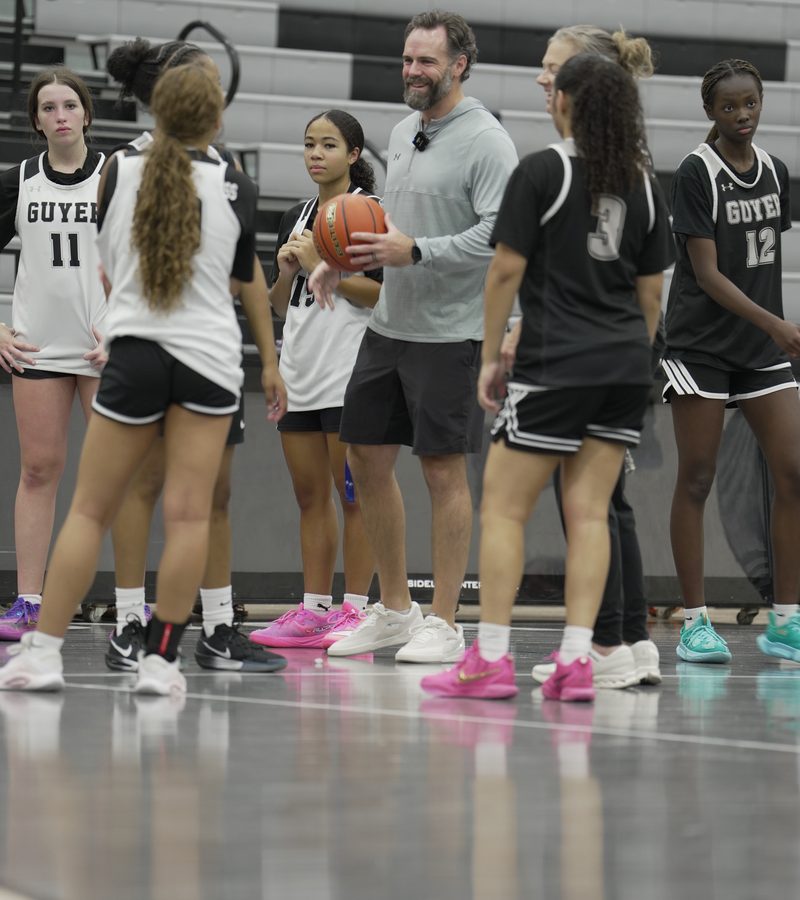 Coach Floyd smiles with a ball in hand as Emily, Keziah and teammates gather around him in the gym.